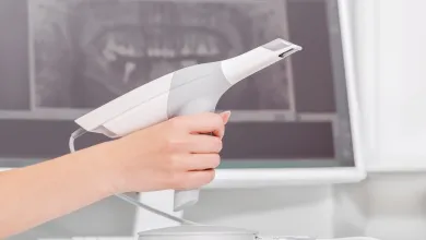 Hand holding a white and gray intraoral scanner, with a dental X-ray on a blurred computer screen in the background.