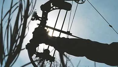 Silhouette of an archer holding a compound bow against a bright sun.