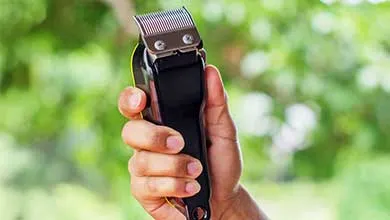 Hand holding black and silver electric hair clippers against a blurred green background.