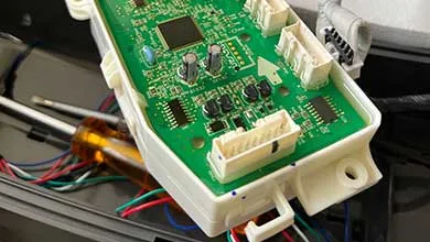 Close-up of a green circuit board in a white housing, with a screwdriver and colored wires blurred in the background.