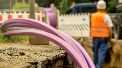 Close-up of purple fiber optic cables in a construction trench with a worker in safety gear blurred in the background.