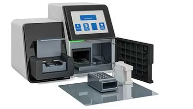 A white and black laboratory device with a digital touchscreen and an open front compartment. A sample tray and cartridge are placed on a gray mat in front of the machine.