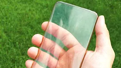 A hand holds a transparent smartphone, showing the palm and green grass clearly through the screen.
