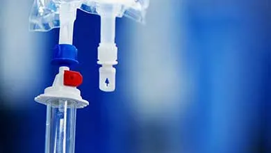 Close-up of an IV infusion bag with drip chamber, connectors, and tubing in a clinical environment.