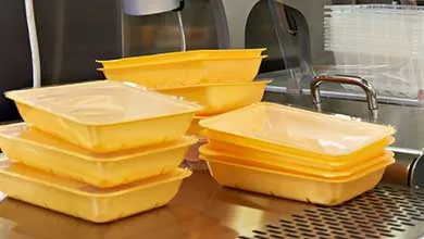 Stacks of yellow rectangular plastic food containers with clear lids on a stainless steel surface, with commercial kitchen equipment in the background.