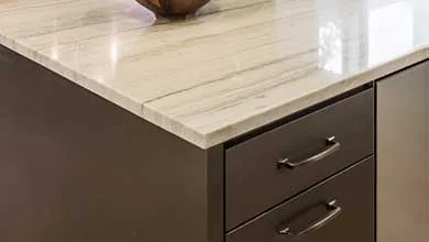 Close-up of a light-colored polished stone countertop with subtle gray veins, resting on dark brown or charcoal cabinetry with two drawers and sleek metallic handles.
