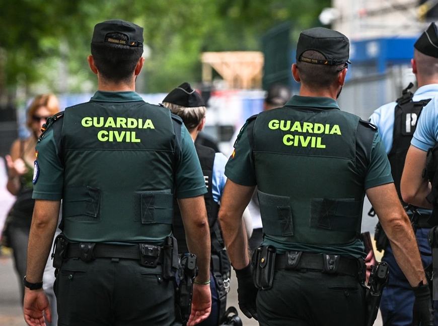 Two Guardia Civil officers, seen from behind, wear dark green uniforms with "GUARDIA CIVIL" in yellow on their backs.