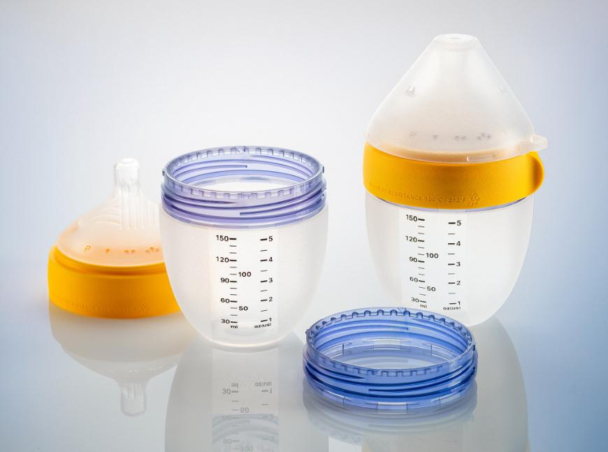 Two baby bottles and their parts on a reflective surface. One bottle is fully assembled with a yellow ring; the other is disassembled, showing a milk-filled container beside its purple and blue rings.