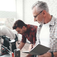 Three men work together in an office; two examine a 3D printer while the third writes in a notebook.
